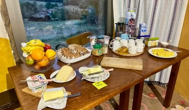 Table du petit déjeuner près d’une fenêtre : fruits, beurre, confitures, pain